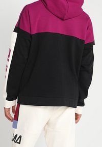 Sweatshirt à capuche noir, avec une partie supérieure magenta et des manches blanches. Présente un imprimé logo avec des accents rouges et bleus sur les manches.
