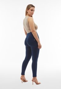 Jeans de tiro alto en azul oscuro con una silueta ajustada, combinados con una blusa sin mangas beige y sandalias de tacón transparente.