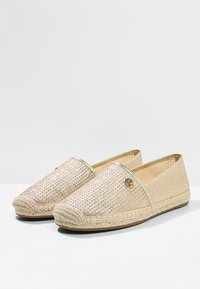 Zapatos tipo slip-on en dorado claro con parte superior tejida, suela trenzada de yute y pequeño logo decorativo en el lateral. Forro interior suave. Diseño plano.