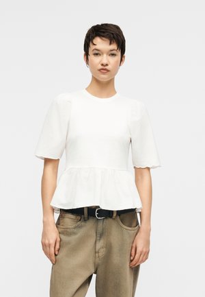 Młoda osoba o krótkich ciemnych włosach, ubrana w białą bluzkę z krótkim rękawem i falistymi krawędziami oraz luźne beżowe spodnie z czarnym paskiem.