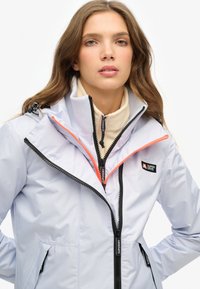 Jasnofioletowa wodoodporna kurtka z czarnymi zamkami i koralowymi akcentami, z wysokim kołnierzem i dwoma bocznymi kieszeniami, wykonana z gładkiego materiału.