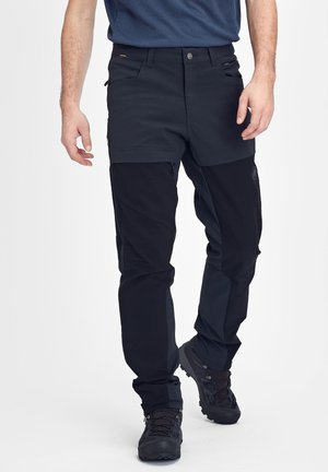 Man draagt zwart-witte hikingbroek en zwarte wandelschoenen, staand tegen een effen lichte achtergrond.