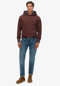 Felpa bordeaux con tasca frontale e cordini, indossata sopra una camicia a quadri; abbinata a jeans blu e stivaletti brown con lacci.