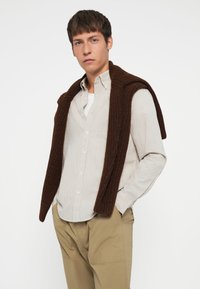 Stripet skjorte i lyse farger, med en mørkebrun strikkegenser drapert over skuldrene, sammen med beige bukser.