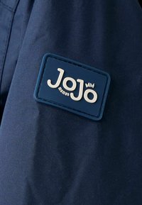 Marineblauer Stoff mit einer strukturierten Oberfläche, versehen mit einem rechteckigen blauen Lederpatch, das mit dem Markennamen "JoJo" in weißen Buchstaben geprägt ist.