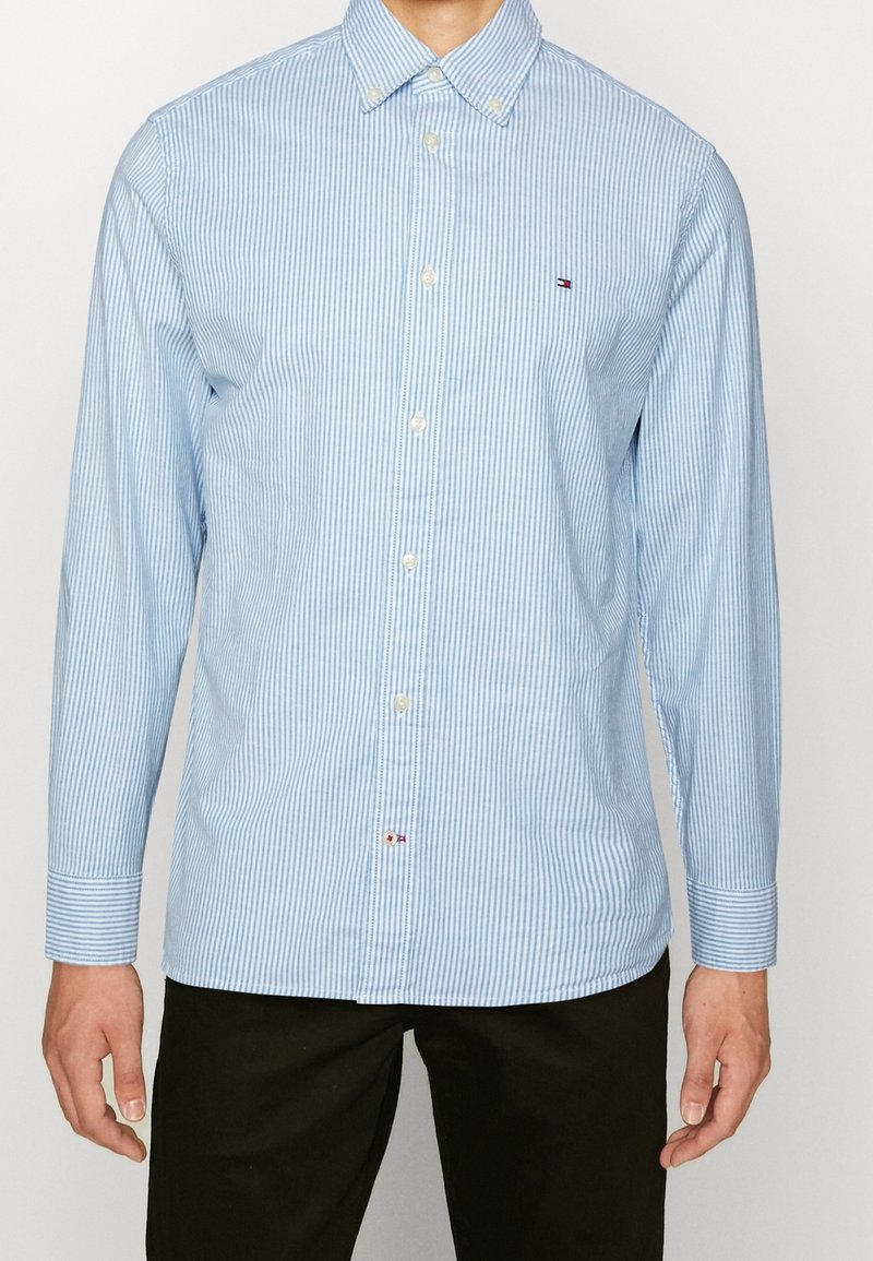 Homme portant une chemise à fines rayures bleu clair et blanc, à boutons, manches longues avec col, et un pantalon noir, debout devant un fond uni.