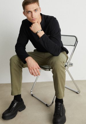 Czarna koszula z długim rękawem, zielone spodnie o zwężanym kroju, czarne sneakersy na platformie i bransoletka. Siedząca na chromowym krześle przy neutralnej ścianie.