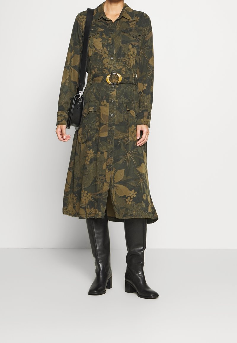 Femme portant une robe verte à fleurs à manches longues avec une ceinture, des bottes noires montantes et un sac noir à bandoulière.