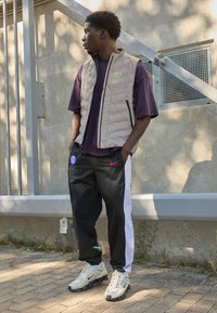 Beige polstret vest over en lilla t-shirt, sorte bukser med hvide striber i siden og lyse sko. Teksturerede materialer med kontrasterende farver.