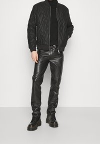 Veste bomber matelassée noire avec des poignets côtelés, associée à un pantalon en cuir noir ajusté et des bottes noires à lacets, sur fond neutre.