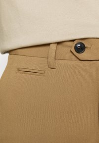 Béžové kalhoty s hladkou texturou, vybavené zapínáním na knoflík a boční kapsou, nošené pod světle béžovým topem.