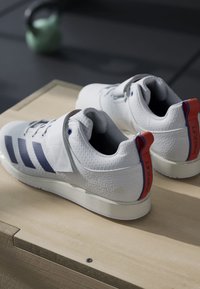 White athletic shoes with three blue stripes, textured upper material, adjustable strap, and red accents on the heel labeled "POWERLIFT."