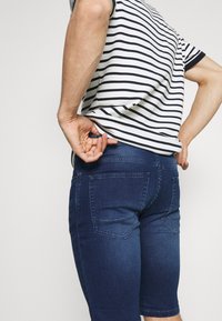 Persona ajustando la cinturilla de unos pantalones cortos de mezclilla azul oscuro, vistiendo una camiseta de manga corta blanca y negra con rayas, frente a un fondo liso.