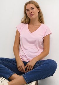 T-shirt rose en V avec des manches courtes retroussées. Tissu lisse, coupe décontractée. Portée avec un jean bleu foncé et dans une pose assise sur un fond neutre.