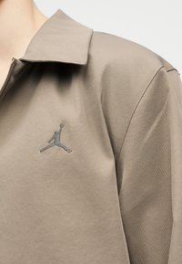 Primo piano di una giacca beige con logo Jumpman ricamato sul petto sinistro, caratterizzata da un colletto e chiusura con zip.