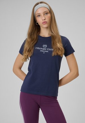 Młoda kobieta z długimi włosami, nosząca szarą opaskę na głowę, granatowy t-shirt "Victory Club" i fioletowe legginsy z białymi bocznymi pasami, pozująca na szarym tle.