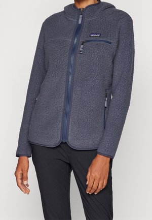 Personne portant une veste polaire bleu à fermeture éclair avec une poche poitrine et le logo Patagonia, associée à un pantalon noir.