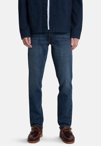 Jeans de mezclilla azul oscuro con corte recto, combinados con una chaqueta navy, sobre un fondo blanco liso. Unos zapatos marrones con cordones completan el look.