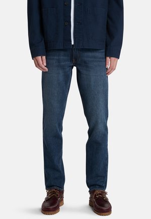 Jeans in denim blu scuro con taglio dritto, abbinati a una giacca blu navy, posti su uno sfondo bianco semplice. Scarpe in pelle marroni con lacci completano il look.