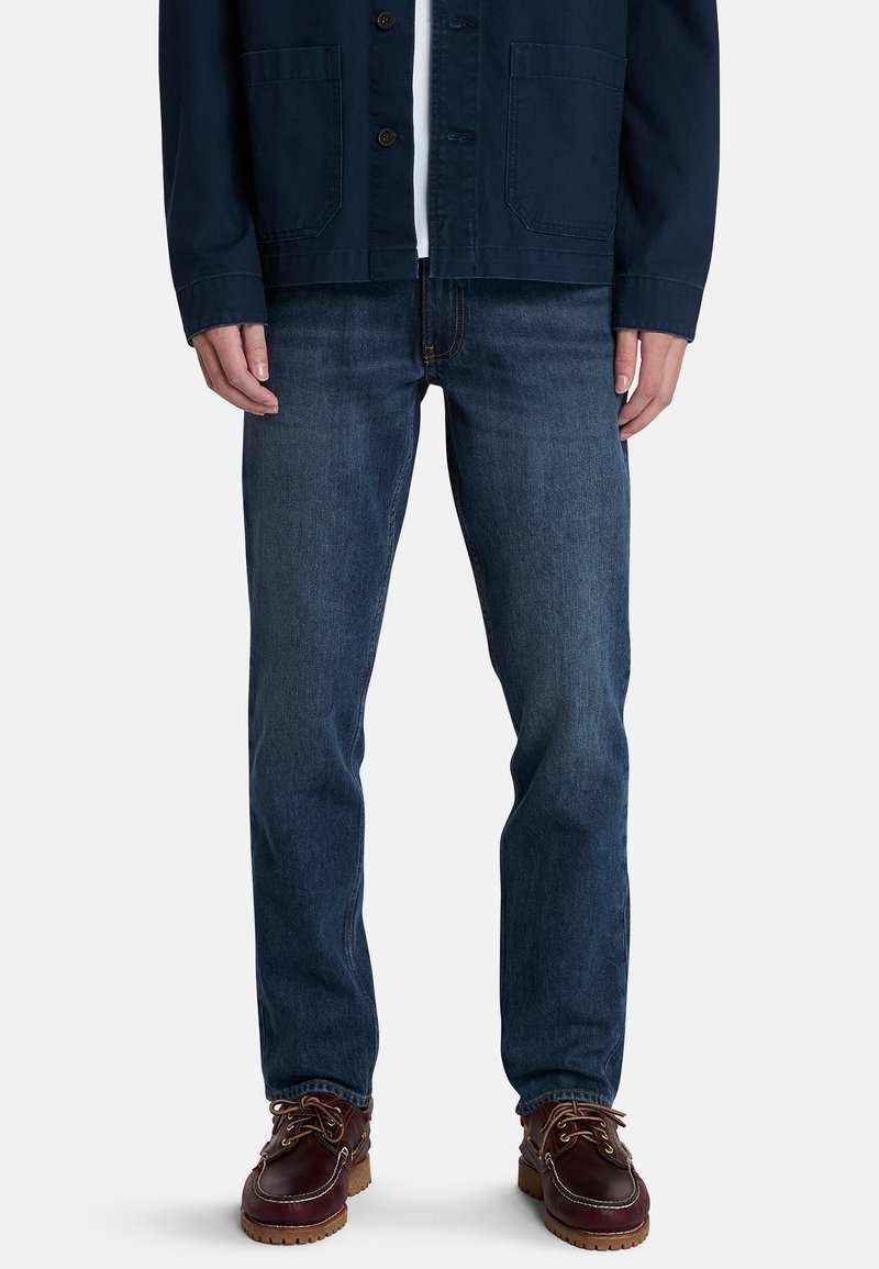 Jeans de mezclilla azul oscuro con corte recto, combinados con una chaqueta navy, sobre un fondo blanco liso. Unos zapatos marrones con cordones completan el look.