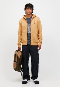 Brun zip-up hoodie, grå grafisk t-shirt, sort cargo bukser, holdende en brun og sort rygsæk, stående mod en hvid baggrund.