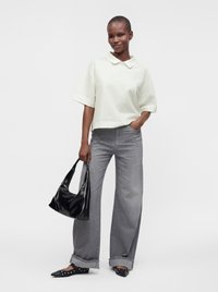 Femme portant un haut blanc texturé à manches courtes, un jean gris large, des ballerines noires cloutées, tenant un sac à main noir, debout devant un fond uni.