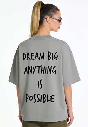 Femme avec queue de cheval blonde portant un t-shirt gris oversize avec le texte "DREAM BIG ANYTHING IS POSSIBLE" dans le dos.