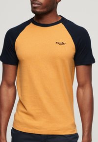 Camiseta de manga corta con un cuerpo amarillo mostaza y mangas raglán azul marino. Presenta un logo sutil en el pecho, tejido suave y un corte estándar.