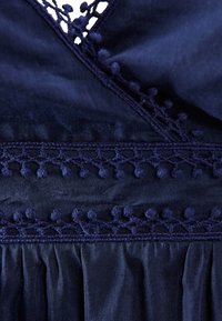 Námorně modrá tkanina s jemným třásňovým lemem a krajkovými detaily kolem výstřihu, s nařasenou texturou na spodní části.
