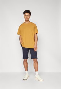 T-shirt a maniche corte color senape con una vestibilità comoda, abbinato a pantaloni corti blu navy. Il modello indossa sneaker bianche e calze bianche a lunghezza crew.