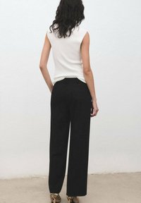 Donna con capelli ondulati scuri che indossa un top bianco senza maniche, pantaloni neri a gamba larga e tacchi alti con motivo pitone, in piedi contro un muro.