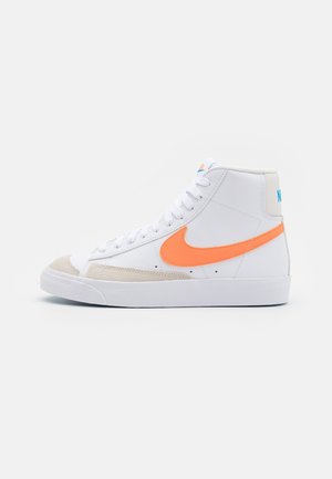 Hvid højtstøvle-sneaker med orange Nike-swoosh, beige tåaccent, hvide snørebånd og blå Nike-tekst på hælen mod en ensfarvet baggrund.