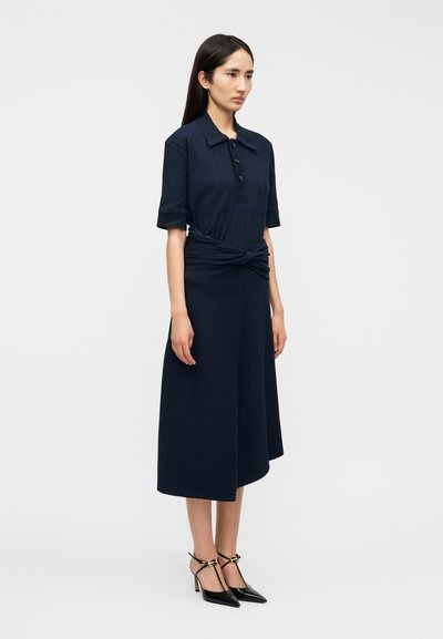 Φόρεμα μήκους έως το γόνατο σ ε ναυτικό μπλε χρώμα, με κολάρο με κουμπιά, κοντά μανίκια και λεπτομέρεια με δετό στη μέση. Φοριέται με μαύρα ψηλοτάκουνα παπούτσια με μυτερή μύτη.