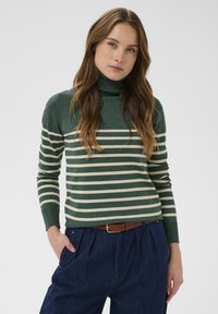 Zeleno prugasta turtleneck pulover s kremasto vodoravnim prugama, rebrastim manžetami in prilegnjenim krojem, v kombinaciji s temno modrimi visokim hlačami.