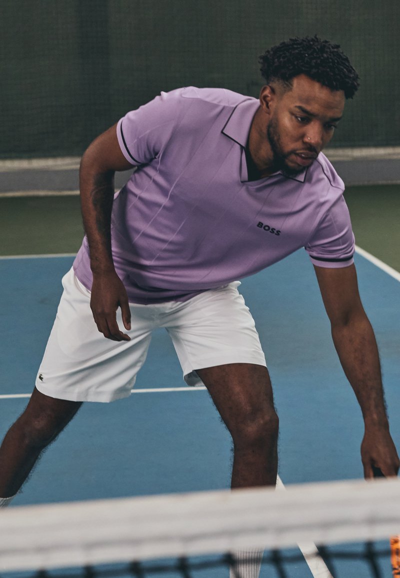 Violet sportinė polo marškinėliai su juodomis detalėmis, trumpomis rankovėmis ir logotipu ant krūtinės, derinami su baltais šortais teniso korte.