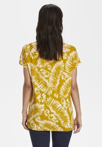 Lyhyet hihnatoppi sinapinkeltaisena, jossa on valkoinen kukkakuosi ja lehtimäinen kuvio. Sujuva kangas, rento istuvuus, kaareva helma.