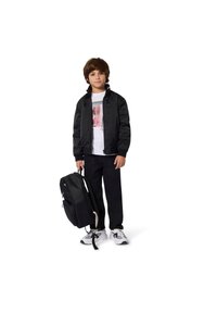 Veste noire matelassée, T-shirt blanc avec impression graphique, pantalon noir et sac à dos noir. Les chaussures sont blanches avec des accents bleus.