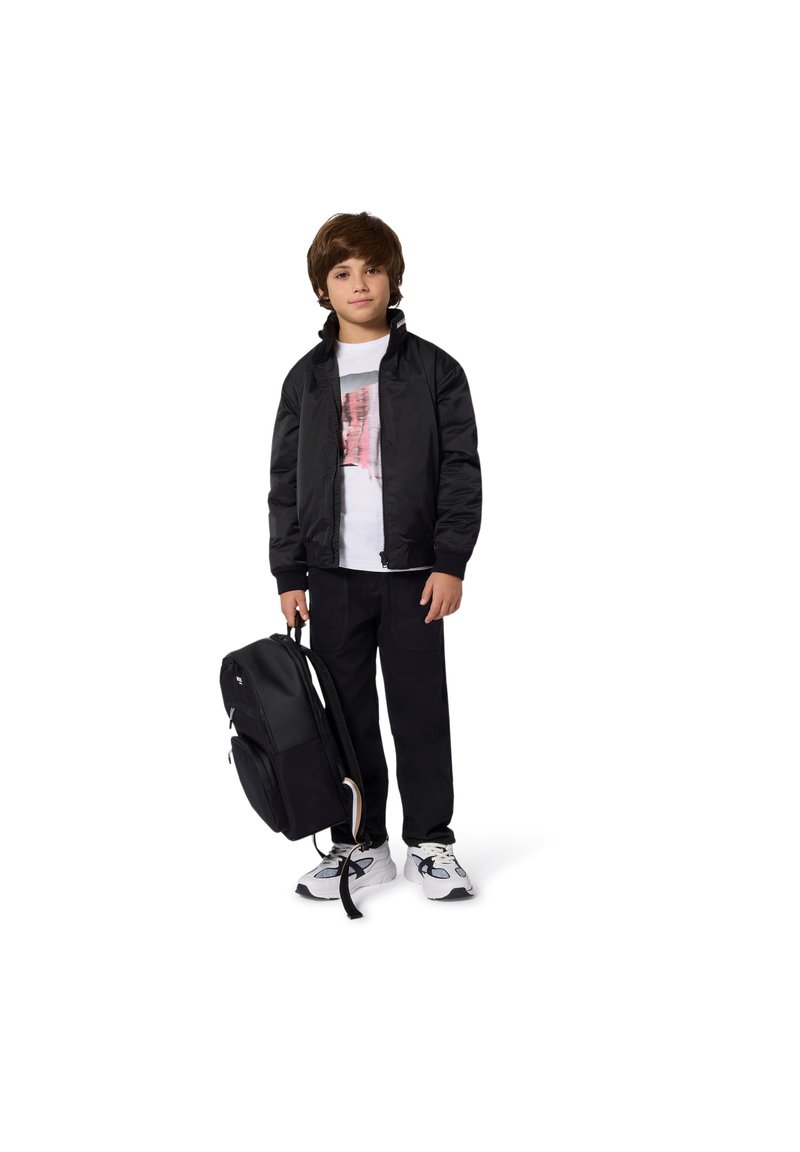 Veste noire matelassée, T-shirt blanc avec impression graphique, pantalon noir et sac à dos noir. Les chaussures sont blanches avec des accents bleus.