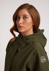 Olivgrön vattentät jacka med hög krage, justerbar huva och logopatch på ärmen. Slät textur, dragkedja/knappstängning.