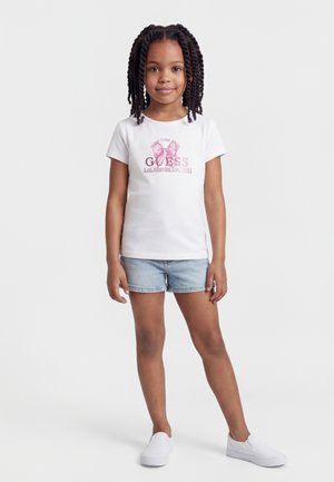 Νεαρή κοπέλα με πλεξούδες, φορώντας λευκό μπλουζάκι με στάμπα, ανοιχτόχρωμα τζιν σορτς και λευκά παπούτσια χωρίς κορδόνια, στέκεται μπροστά σε απλό λευκό φόντο.