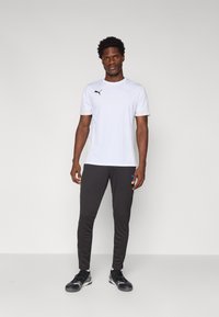 Balta sportinė marškinė su juodu Puma logotipu, derinami su tamsiai pilkais siaurais treningais, kurių kairėje šlaunyje yra mažas mėlynas logotipas.