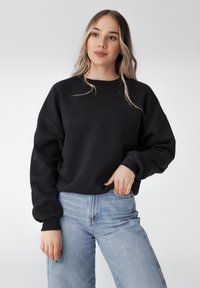 Sweatshirt en coton noir avec un col rond et des manches surdimensionnées. Présente un détail de logo subtil près du col. Assorti avec un jean bleu clair.