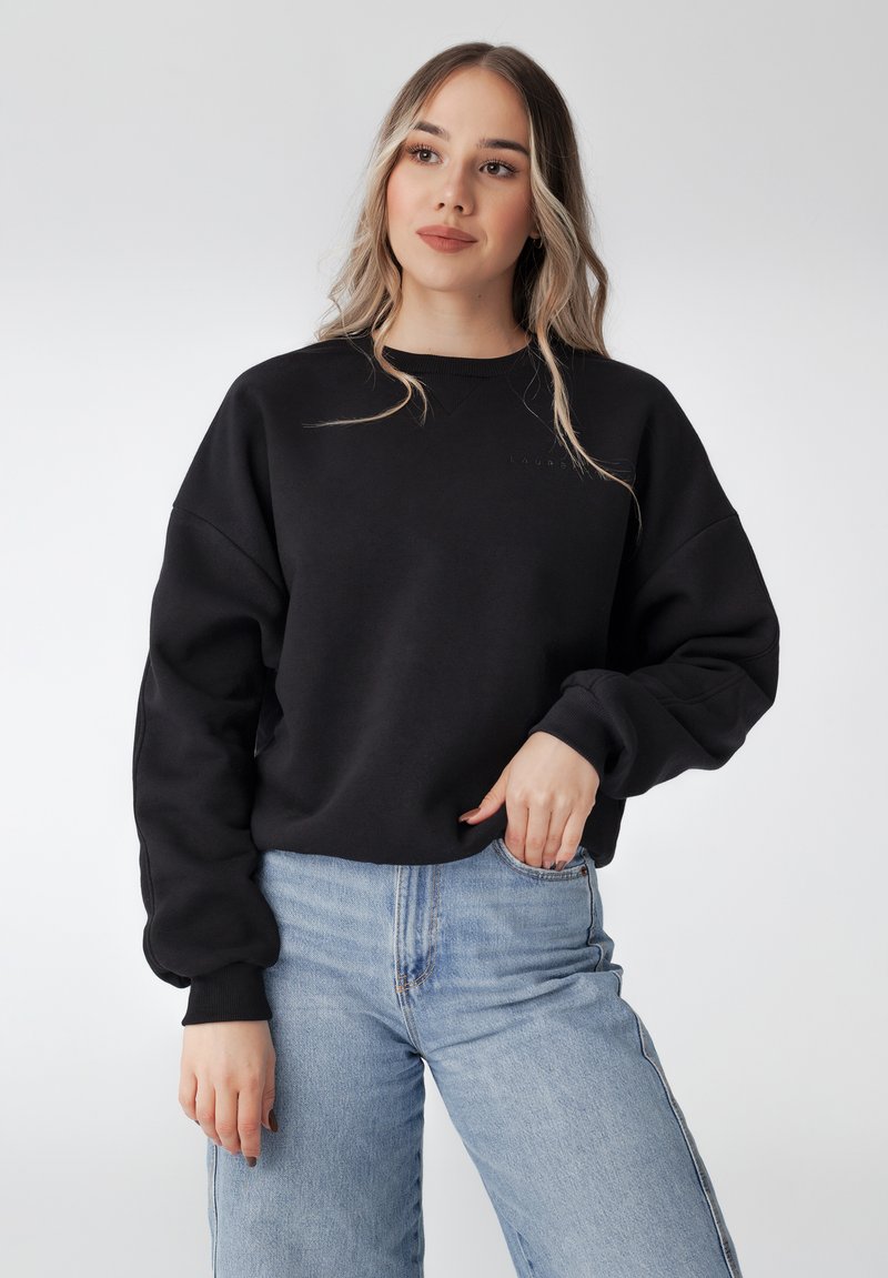 Sweatshirt en coton noir avec un col rond et des manches surdimensionnées. Présente un détail de logo subtil près du col. Assorti avec un jean bleu clair.