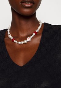 Femme portant un haut noir texturé à col en V et un collier de perles avec des perles rouges et une perle blanche en forme de cœur avec des points rouges.