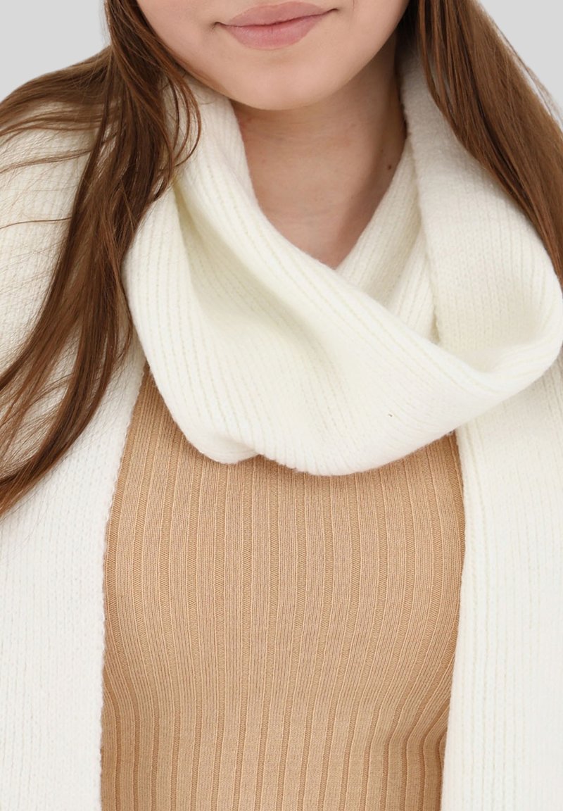 Écharpe en tricot côtelé crème enroulée autour du cou, superposée à un haut beige côtelé ajusté. Texture douce et palette de couleurs neutres.