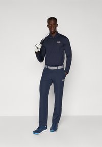 Mornarski golf komplet z dolgimi rokavi, vzorčastim pasom in usklajenimi hlačami. Model drži golf palico in ima belo rokavico.