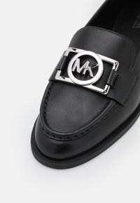 Mocassin en cuir noir avec boucle logo Michael Kors argentée sur le dessus, couture détaillée autour de la pointe, et semelle lisse visible.
