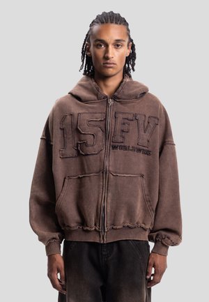 Giovane uomo con capelli intrecciati che indossa una felpa marrone con zip e scritta "15 FV Worldwide", in piedi davanti a uno sfondo grigio uniforme.