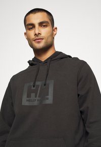 Hoodie noir en tissu doux avec un logo noir bien visible sur la poitrine. Comprend une capuche avec cordon de serrage et des poignets côtelés.