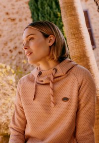 Mladá žena s krátkymi blond vlasmi, oblečená v svetlo hnedom mikinovom pulóvri s bodkami, stojaca vonku pri strome a kamennej stene v jemnom slnečnom svetle.